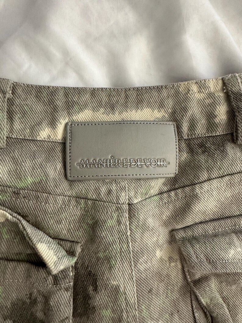 May include: A pair of grey denim pants featuring a mottled, tie-dye style pattern with subtle hints of light green. A rectangular grey leather-like patch is stitched to the back waistband, embossed with the text MANIERE DE VOIR in capital letters.