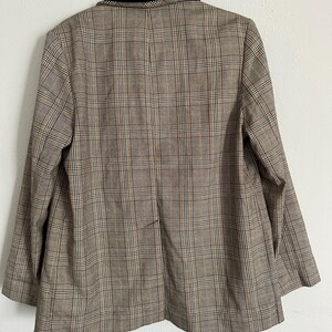 May include: A brown plaid blazer with a black velvet collar, hanging against a white wall. The blazer features a classic plaid pattern in shades of brown, beige, and a hint of red. The back of the blazer is visible, showcasing its tailored design.