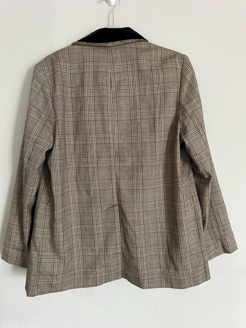 May include: A brown plaid blazer with a black velvet collar, hanging against a white wall. The blazer features a classic plaid pattern in shades of brown, beige, and a hint of red. The back of the blazer is visible, showcasing its tailored design.