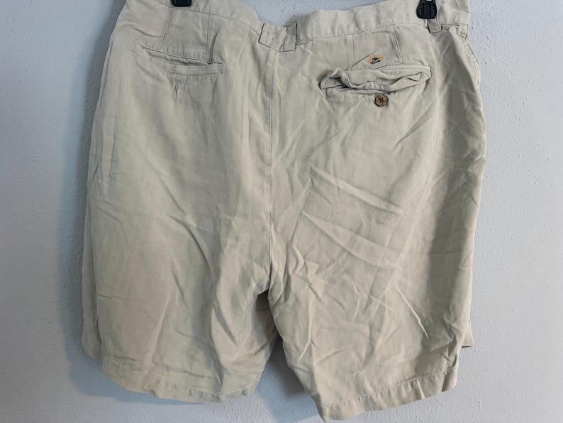 May include: Beige shorts with a buttoned back pocket and two front pockets. The shorts are made of a lightweight fabric and have a relaxed fit. The shorts are hanging on a black hanger.