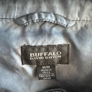 K&ouml;nnte beinhalten: Nahaufnahme einer dunkelblauen Jacke mit einem Etikett mit der Aufschrift "BUFFALO DAVID BITTON". Auf dem Etikett steht auch "M/M Made in China". Die Jacke h&auml;ngt an einem wei&szlig;en Kleiderb&uuml;gel.