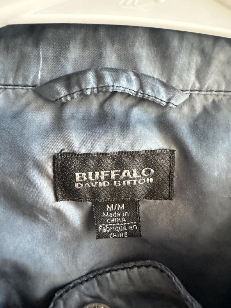 K&ouml;nnte beinhalten: Nahaufnahme einer dunkelblauen Jacke mit einem Etikett mit der Aufschrift "BUFFALO DAVID BITTON". Auf dem Etikett steht auch "M/M Made in China". Die Jacke h&auml;ngt an einem wei&szlig;en Kleiderb&uuml;gel.