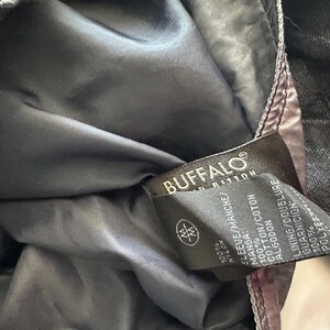K&ouml;nnte beinhalten: Nahaufnahme des Innenraums einer schwarzen Jacke mit dunkelgrauem Futter. Ein Etikett mit der Aufschrift "BUFFALO" enth&auml;lt Angaben zu den Materialien: 100 % Nylon f&uuml;r den &Auml;rmel, 100 % Baumwolle und 100 % Polyester.