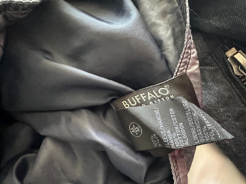 K&ouml;nnte beinhalten: Nahaufnahme des Innenraums einer schwarzen Jacke mit dunkelgrauem Futter. Ein Etikett mit der Aufschrift "BUFFALO" enth&auml;lt Angaben zu den Materialien: 100 % Nylon f&uuml;r den &Auml;rmel, 100 % Baumwolle und 100 % Polyester.