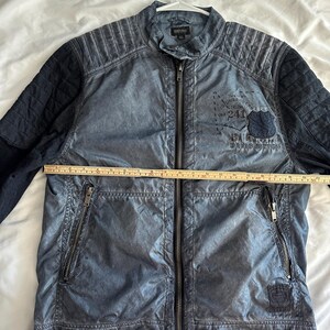 K&ouml;nnte beinhalten: Blaue Jacke mit durchgehendem Rei&szlig;verschluss, zwei Rei&szlig;verschlusstaschen und einem Steppmuster an Schultern und &Auml;rmeln. Die Jacke hat einen Aufn&auml;her mit Text und einem Schilddesign. Die Jacke misst ca. 61 cm quer &uuml;ber die Brust.