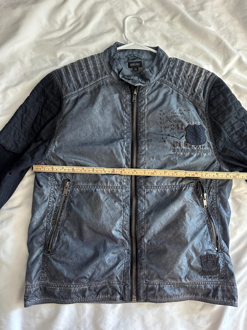 K&ouml;nnte beinhalten: Blaue Jacke mit durchgehendem Rei&szlig;verschluss, zwei Rei&szlig;verschlusstaschen und einem Steppmuster an Schultern und &Auml;rmeln. Die Jacke hat einen Aufn&auml;her mit Text und einem Schilddesign. Die Jacke misst ca. 61 cm quer &uuml;ber die Brust.