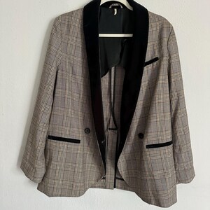 May include: A plaid blazer with a black velvet collar and trim, hanging on a white hanger. The blazer has a double-breasted design with two black buttons and two front pockets. The plaid pattern includes shades of brown, beige, and black.