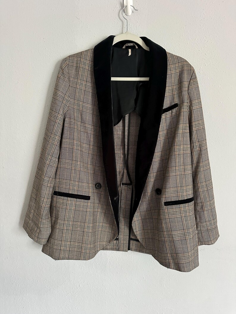 May include: A plaid blazer with a black velvet collar and trim, hanging on a white hanger. The blazer has a double-breasted design with two black buttons and two front pockets. The plaid pattern includes shades of brown, beige, and black.