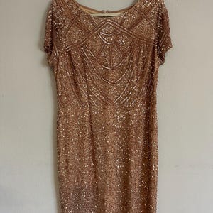 Puede incluir: Un vestido hasta la rodilla en color oro rosa con mangas cortas, cuello redondo y un intrincado patr&oacute;n geom&eacute;trico de lentejuelas y cuentas que cubre toda la superficie. La prenda se muestra en una percha blanca.