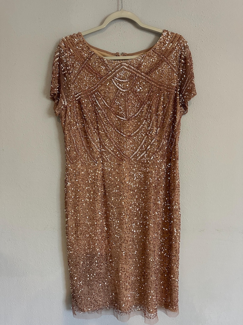 Puede incluir: Un vestido hasta la rodilla en color oro rosa con mangas cortas, cuello redondo y un intrincado patr&oacute;n geom&eacute;trico de lentejuelas y cuentas que cubre toda la superficie. La prenda se muestra en una percha blanca.