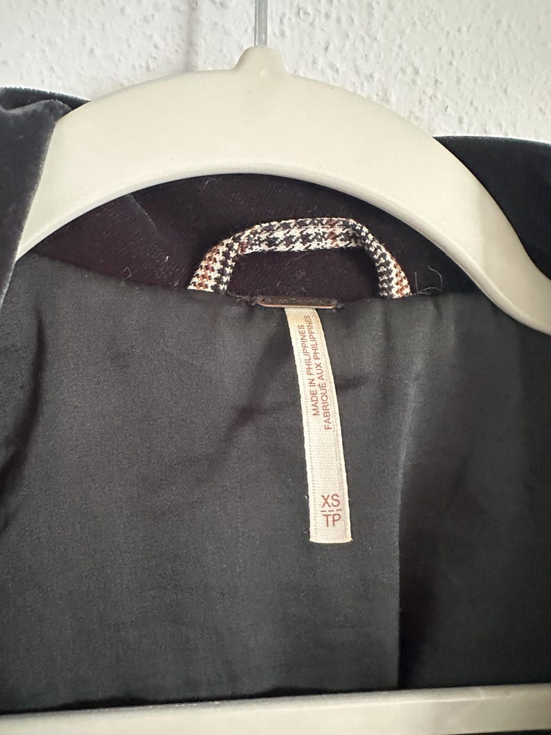 May include: Close-up of a dark gray velvet jacket hanging on a white hanger. The jacket's interior lining is black, with a brown and white patterned collar detail. A tag reads "MADE IN PHILIPPINES" and "XS TP".