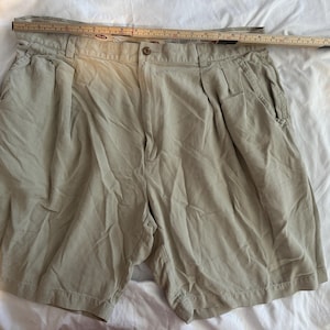 May include: Beige shorts with pleats, a button fastening, and pockets. A wooden ruler is placed across the top of the shorts. The shorts are made of a lightweight fabric and are laid out on a white surface. The shorts are a neutral colour.