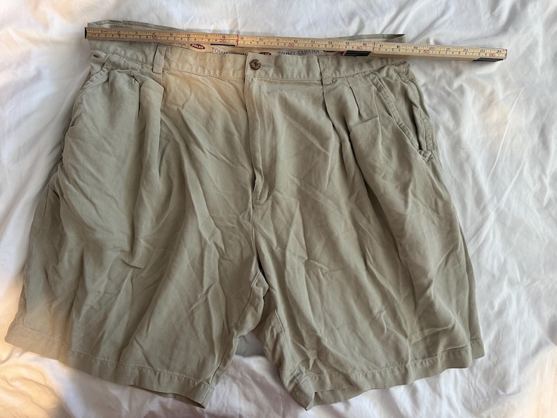 May include: Beige shorts with pleats, a button fastening, and pockets. A wooden ruler is placed across the top of the shorts. The shorts are made of a lightweight fabric and are laid out on a white surface. The shorts are a neutral colour.