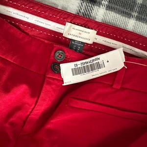 May include: Close-up of red pants with two dark buttons and a tag. The tag reads "the essential SLIM by ANTHROPOLOGIE". The pants have a smooth texture and a small pocket. The tag also has a barcode and the text "APP23Y46452-03".