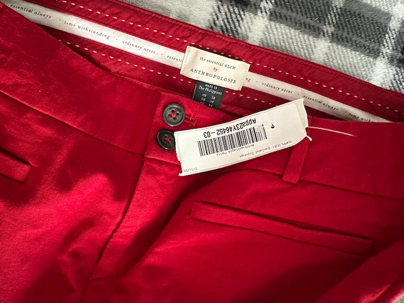 May include: Close-up of red pants with two dark buttons and a tag. The tag reads "the essential SLIM by ANTHROPOLOGIE". The pants have a smooth texture and a small pocket. The tag also has a barcode and the text "APP23Y46452-03".