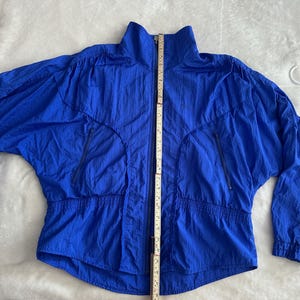 May include: A bright blue jacket with a high collar and a full-length zip. The jacket has two zipped front pockets and an elasticated waistband. The jacket is approximately 76 cm long, measured from the top of the collar to the bottom hem.