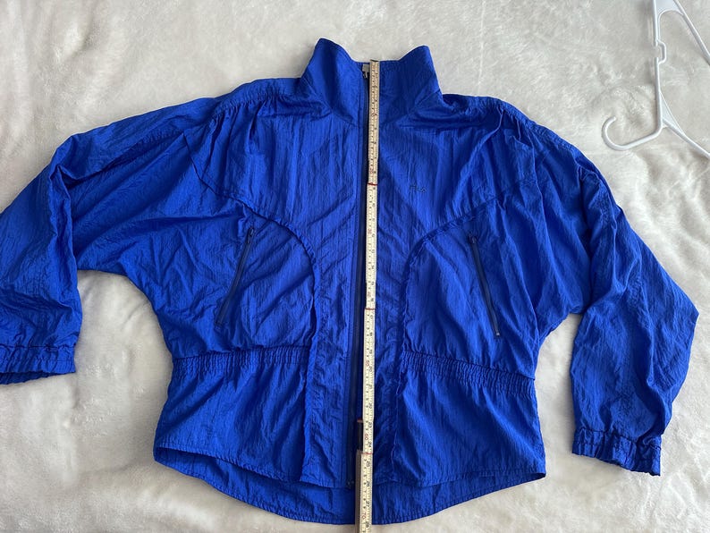 May include: A bright blue jacket with a high collar and a full-length zip. The jacket has two zipped front pockets and an elasticated waistband. The jacket is approximately 76 cm long, measured from the top of the collar to the bottom hem.