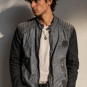 K&ouml;nnte beinhalten: Jeansjacke in verwaschenem Blau und Schwarz mit Rei&szlig;verschluss und kleinem Aufn&auml;her mit Text. Die Jacke hat ein geripptes Design an den Schultern. Das Model tr&auml;gt ein wei&szlig;es T-Shirt und eine silberne Halskette.