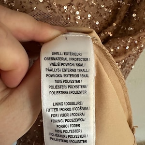 Puede incluir: Una etiqueta de cuidado de tela blanca unida a una prenda cubierta de lentejuelas de color oro rosa. El texto de la etiqueta dice: SHELL / EXT&Eacute;RIEUR / OBERMATERIAL / PROTECTOR / VNĚJ&Scaron;&Iacute; POVRCH / SKAL / P&Auml;&Auml;LLYS / ESTERNO / SKALL / POWŁOKA / EXTERIOR / SKAL 100% POLYESTER.