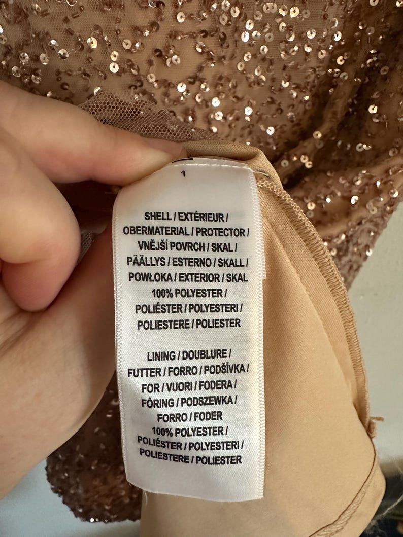 Puede incluir: Una etiqueta de cuidado de tela blanca unida a una prenda cubierta de lentejuelas de color oro rosa. El texto de la etiqueta dice: SHELL / EXT&Eacute;RIEUR / OBERMATERIAL / PROTECTOR / VNĚJ&Scaron;&Iacute; POVRCH / SKAL / P&Auml;&Auml;LLYS / ESTERNO / SKALL / POWŁOKA / EXTERIOR / SKAL 100% POLYESTER.