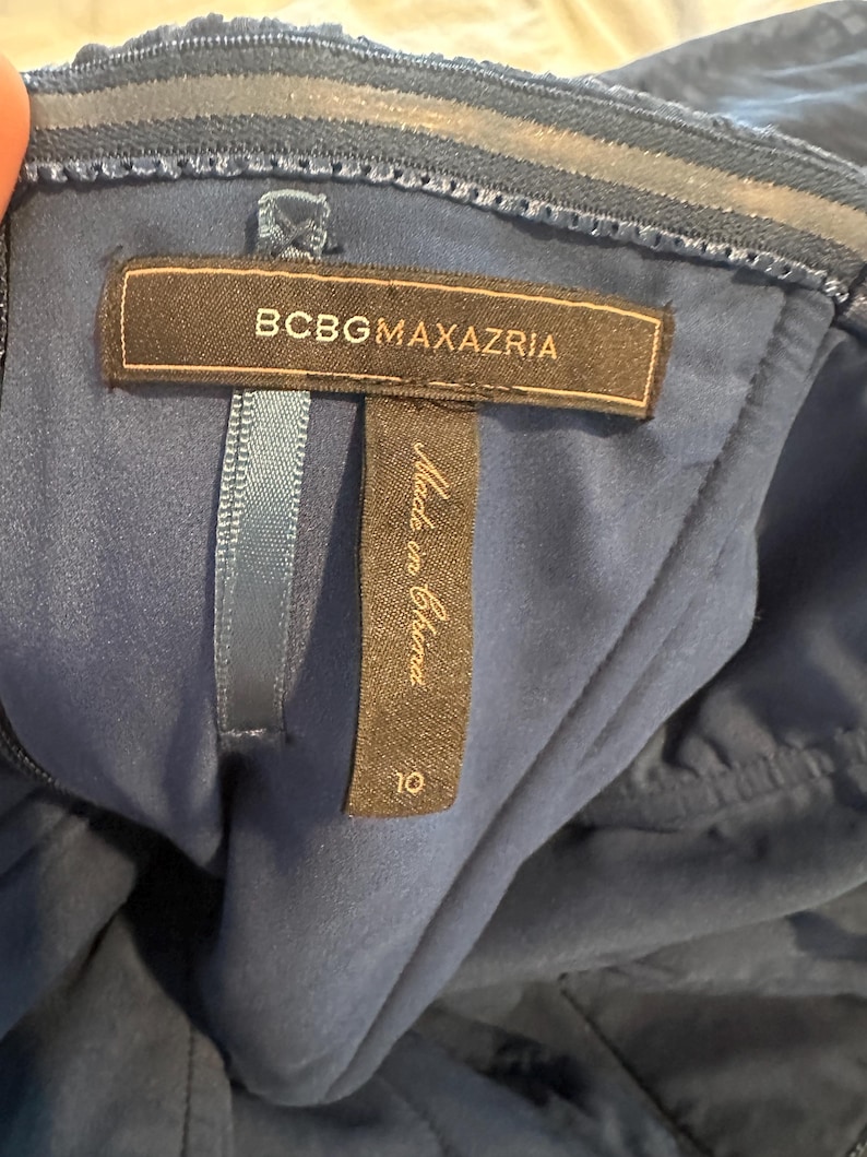 Peut inclure: Gros plan d'une robe bleu marine avec une &eacute;tiquette "BCBGMAXAZRIA". L'&eacute;tiquette indique &eacute;galement "Made in China" et une taille 10. La robe a un tissu textur&eacute; et un design sans bretelles.