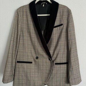May include: A plaid blazer with a black velvet collar and trim. The double-breasted jacket features a classic pattern in shades of brown, beige, and black. The blazer is hanging on a white hanger against a white background.