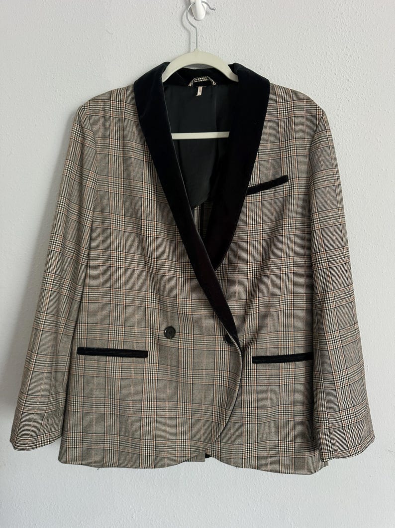 May include: A plaid blazer with a black velvet collar and trim. The double-breasted jacket features a classic pattern in shades of brown, beige, and black. The blazer is hanging on a white hanger against a white background.