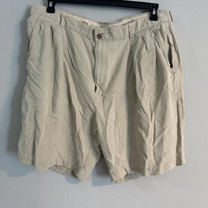 May include: Beige shorts with a button fastening and pockets, hanging on a black hanger. The shorts are against a pale blue wall. The shorts appear to be made of a lightweight material.