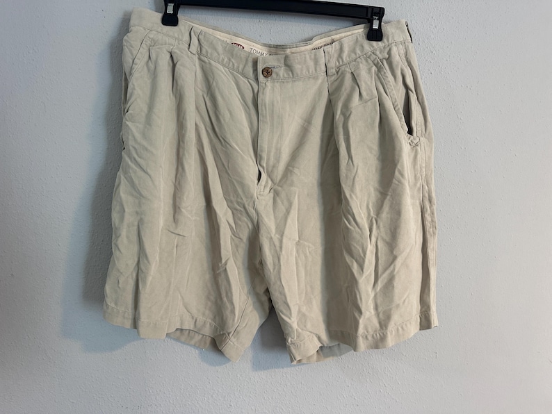 May include: Beige shorts with a button fastening and pockets, hanging on a black hanger. The shorts are against a pale blue wall. The shorts appear to be made of a lightweight material.