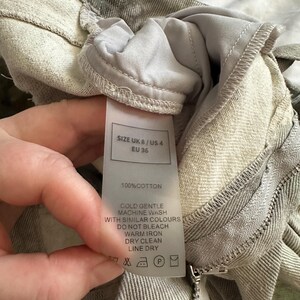 May include: A pair of light gray cotton trousers featuring a visible care label that states SIZE UK 8 / US 4 / EU 36, 100% COTTON, and provides instructions for cold gentle machine wash, do not bleach, warm iron, dry clean, and line dry.
