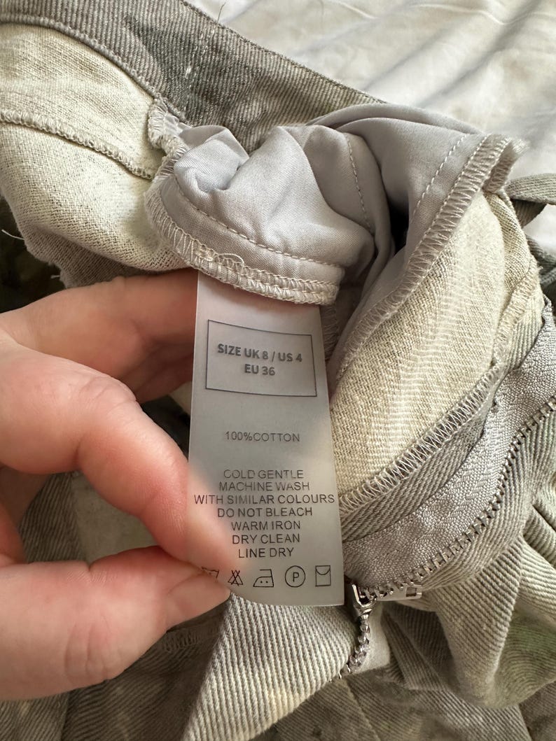 May include: A pair of light gray cotton trousers featuring a visible care label that states SIZE UK 8 / US 4 / EU 36, 100% COTTON, and provides instructions for cold gentle machine wash, do not bleach, warm iron, dry clean, and line dry.