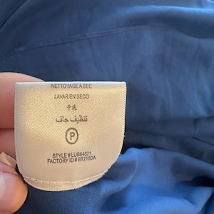 Peut inclure: Une &eacute;tiquette de v&ecirc;tement blanche avec le texte "NETTOYAGE ASEC", "LAVAR EN SECO", "干洗", et "تنظيف جاف". L'&eacute;tiquette comprend &eacute;galement le num&eacute;ro de style "LUB64621" et l'ID d'usine "BT216DA". L'&eacute;tiquette est contre un tissu bleu.