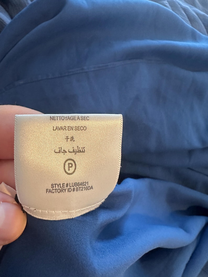 Peut inclure: Une &eacute;tiquette de v&ecirc;tement blanche avec le texte "NETTOYAGE ASEC", "LAVAR EN SECO", "干洗", et "تنظيف جاف". L'&eacute;tiquette comprend &eacute;galement le num&eacute;ro de style "LUB64621" et l'ID d'usine "BT216DA". L'&eacute;tiquette est contre un tissu bleu.