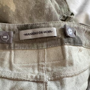 May include: A pair of olive green corduroy pants featuring a waistband with adjustable elastic side tabs and buttons. A white fabric label is sewn onto the center back of the waistband with the black text MANI&Egrave;RE DE VOIR printed on it.