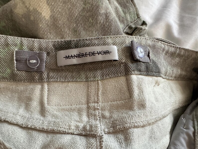May include: A pair of olive green corduroy pants featuring a waistband with adjustable elastic side tabs and buttons. A white fabric label is sewn onto the center back of the waistband with the black text MANI&Egrave;RE DE VOIR printed on it.