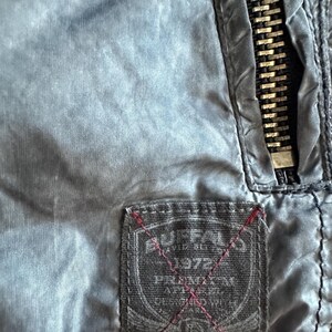 K&ouml;nnte beinhalten: Nahaufnahme einer blau-grauen Jacke mit einem Metallrei&szlig;verschluss und einem rechteckigen Etikett. Das Etikett tr&auml;gt den Text "BUFFALO 1972" und andere Texte mit roten Nahtdetails. Die Jacke hat eine leicht zerknitterte Textur.