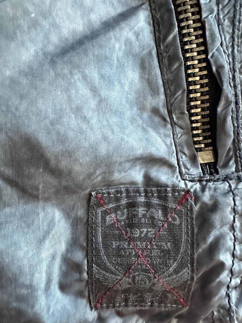 K&ouml;nnte beinhalten: Nahaufnahme einer blau-grauen Jacke mit einem Metallrei&szlig;verschluss und einem rechteckigen Etikett. Das Etikett tr&auml;gt den Text "BUFFALO 1972" und andere Texte mit roten Nahtdetails. Die Jacke hat eine leicht zerknitterte Textur.