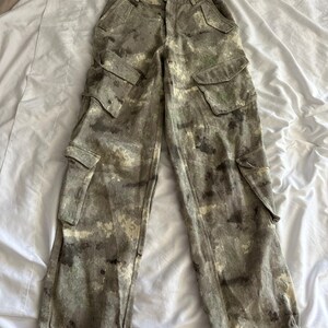 May include: High-waisted cargo pants featuring a mottled camouflage pattern in shades of olive green, brown, and cream. These pants include two front hip pockets, two large side cargo pockets with flaps, and adjustable ties at the bottom hems.