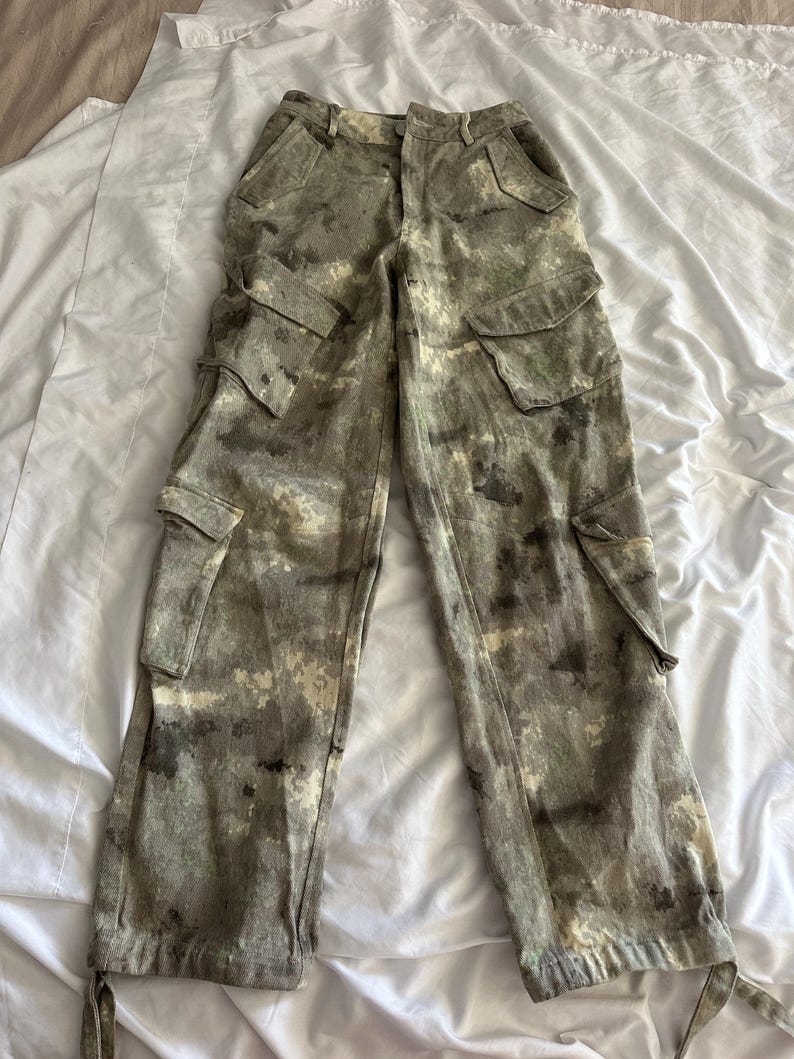 May include: High-waisted cargo pants featuring a mottled camouflage pattern in shades of olive green, brown, and cream. These pants include two front hip pockets, two large side cargo pockets with flaps, and adjustable ties at the bottom hems.