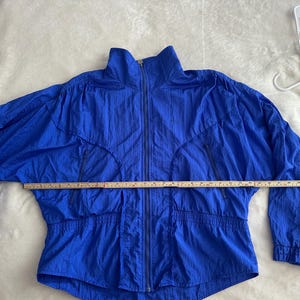 May include: A bright blue vintage jacket with a full zip, two front pockets, and a high collar. The jacket is made of a lightweight, crinkled fabric. A measuring tape is laid across the jacket, indicating a length of approximately 76 cm.