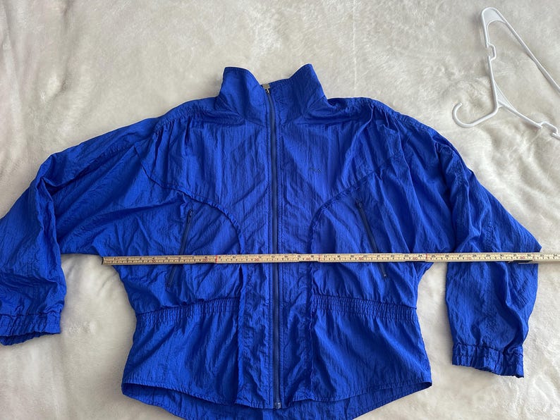 May include: A bright blue vintage jacket with a full zip, two front pockets, and a high collar. The jacket is made of a lightweight, crinkled fabric. A measuring tape is laid across the jacket, indicating a length of approximately 76 cm.