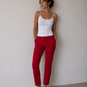 May include: A white spaghetti strap tank top paired with bright red pants. The pants have a tailored fit and a button closure. Beige slip-on shoes complete the outfit. The model is standing in front of a neutral wall.