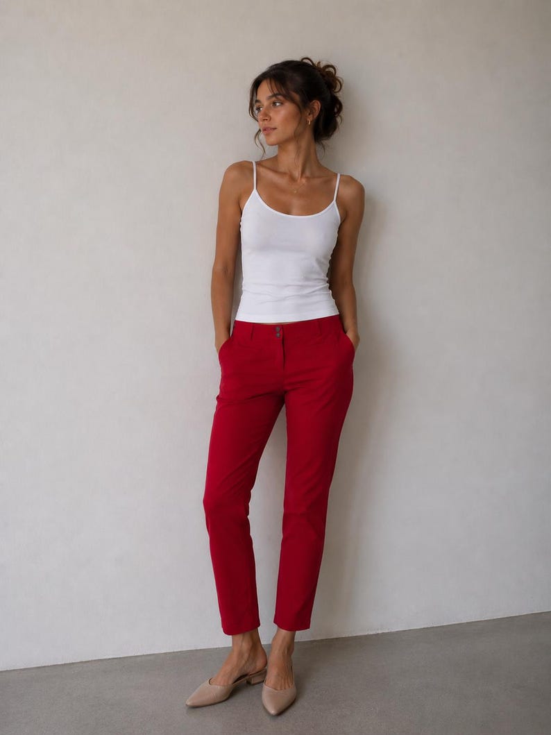 May include: A white spaghetti strap tank top paired with bright red pants. The pants have a tailored fit and a button closure. Beige slip-on shoes complete the outfit. The model is standing in front of a neutral wall.