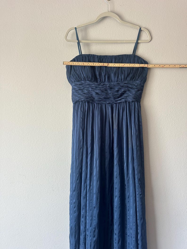 Peut inclure: Une robe de soir&eacute;e bleu marine &agrave; fines bretelles, suspendue &agrave; un cintre blanc. La robe pr&eacute;sente un corsage fronc&eacute; et une jupe fluide &agrave; rayures verticales. Une r&egrave;gle en bois est plac&eacute;e sur le corsage, mesurant environ 76 cm.