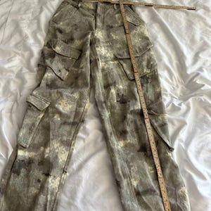 May include: Cargo pants in a mottled gray and white camouflage pattern featuring two large side pockets with flaps and two smaller pockets on the thighs. A wooden folding ruler is placed across the waist and down the leg for measurement.