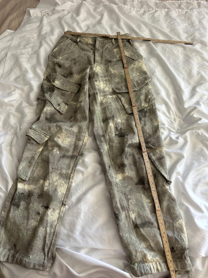 May include: Cargo pants in a mottled gray and white camouflage pattern featuring two large side pockets with flaps and two smaller pockets on the thighs. A wooden folding ruler is placed across the waist and down the leg for measurement.