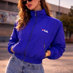 May include: A bright blue Fila jacket with a white logo, featuring a high collar and a zip-up front. The jacket is paired with light-wash denim jeans. The model is wearing black sunglasses and gold hoop earrings.