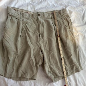May include: Khaki-coloured shorts with a button closure and pleats. The shorts have pockets and a measuring tape is placed next to them. The shorts are made by Tommy Bahama.