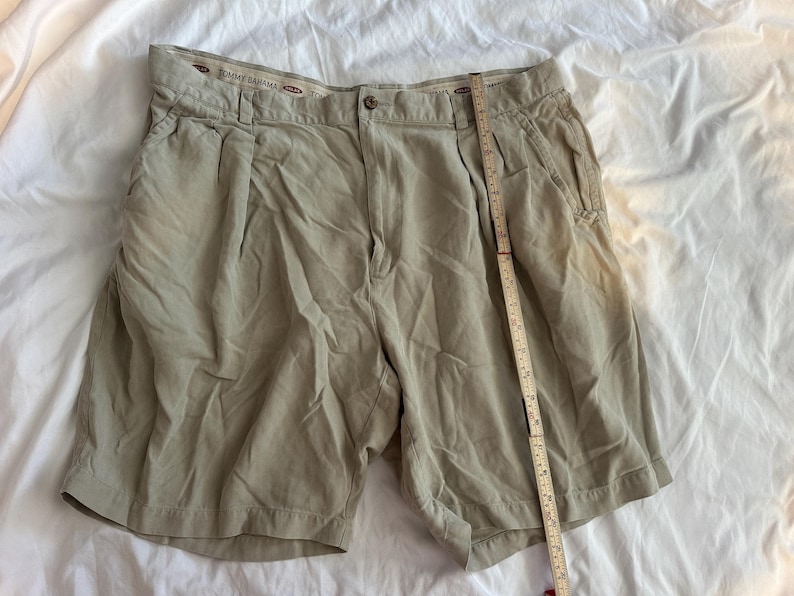 May include: Khaki-coloured shorts with a button closure and pleats. The shorts have pockets and a measuring tape is placed next to them. The shorts are made by Tommy Bahama.