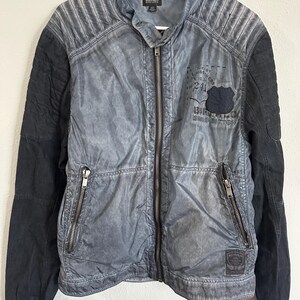 K&ouml;nnte beinhalten: Blau-schwarze Jacke mit Rei&szlig;verschluss. Die Jacke hat ein gestepptes Design an den Schultern und schwarze &Auml;rmel. Ein Aufn&auml;her mit Text und einem Schilddesign ist sichtbar. Die Jacke h&auml;ngt an einem wei&szlig;en Kleiderb&uuml;gel.